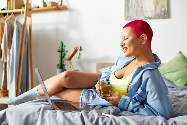 The young woman lounges comfortably on her bed, sipping her drink and smiling cheerfully. — Stock Photo