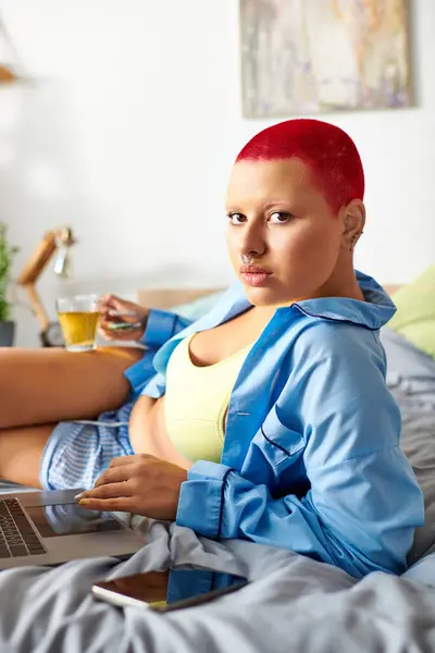 A young woman relaxes at home, sipping tea and using her laptop in comfortable clothing. — Stock Photo