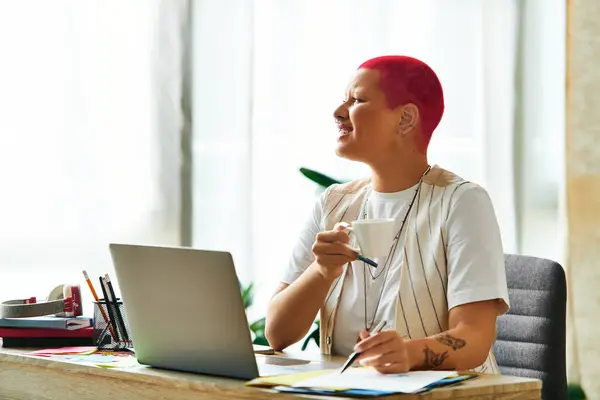 A cheerful young woman sips coffee at her desk, immersed in her creative home workspace. — Stock Photo