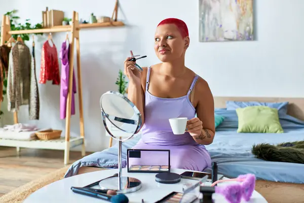 A young woman sits at her table, sipping coffee and applying makeup in a bright, inviting space. — Stock Photo