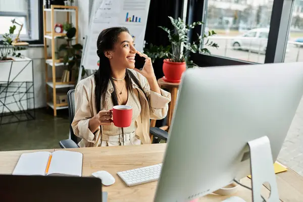 A young woman smiles during a call, holding a red cup, amidst a vibrant office environment. — Stock Photo