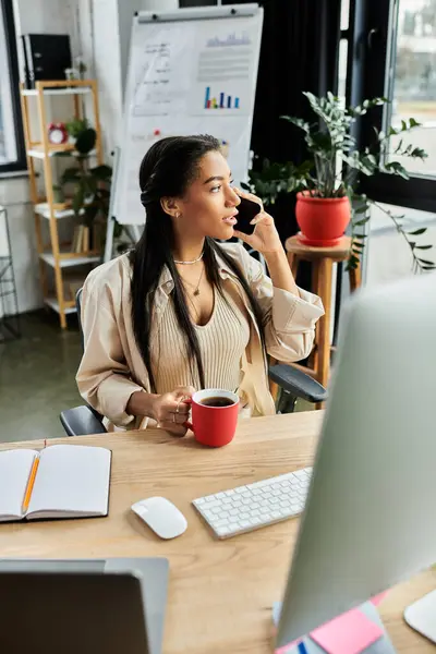 Una joven habla por teléfono, sentada en su escritorio con una taza de café, rodeada de vegetación. - foto de stock