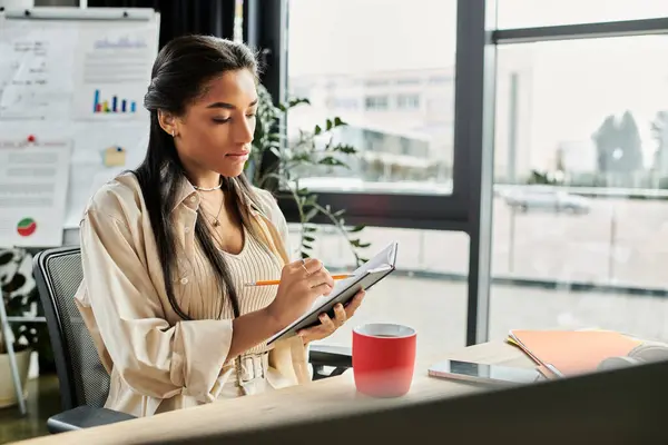 A focused young woman writes notes while sitting at her office desk with large windows. — Stock Photo