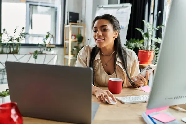A young woman smiles while multitasking on her laptop and phone in a bright space. — Stock Photo