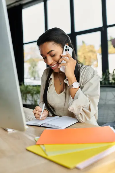 Una joven discute el trabajo por teléfono mientras escribe notas en su escritorio. - foto de stock