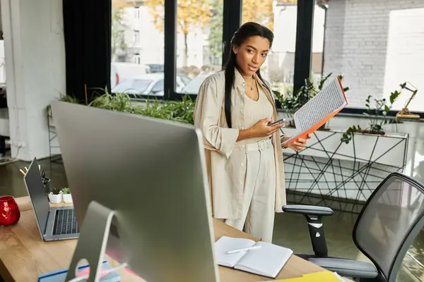 A young woman stands in a stylish office, holding documents and a smartphone, focused on her work. — Stock Photo