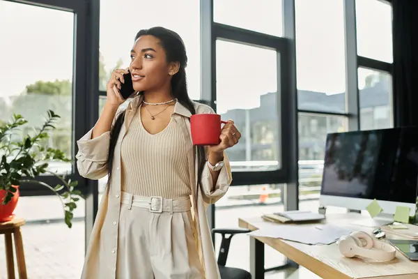A young professional woman speaks on the phone while holding a red cup in an airy office. — Stock Photo