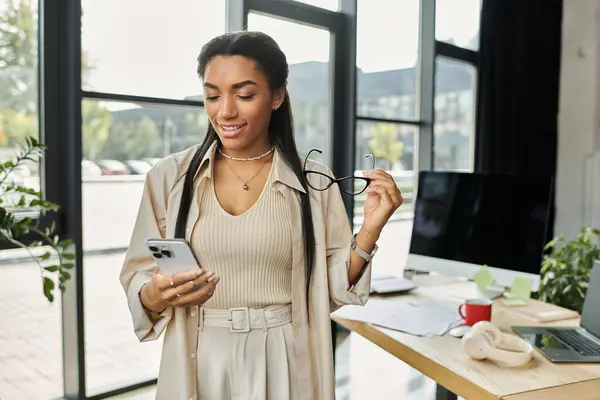 A stylish young woman smiles while looking at her phone in a bright office space. — Stock Photo