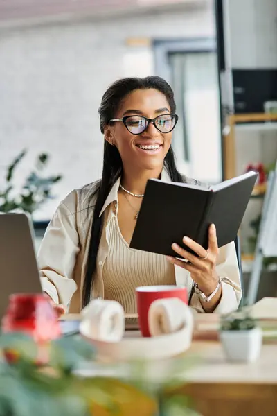 A young professional enthusiastically reads a book at her desk, sipping coffee and smiling. — Stock Photo