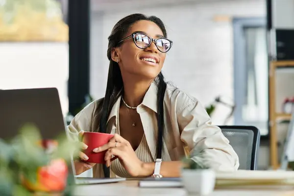 Eine junge schöne Frau lächelt und nippt an ihrem Schreibtisch in einem modernen Büroumfeld am Kaffee.. — Stockfoto