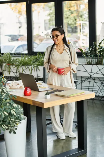 A young woman in business attire smiles while using her laptop in a bright office. — Stock Photo