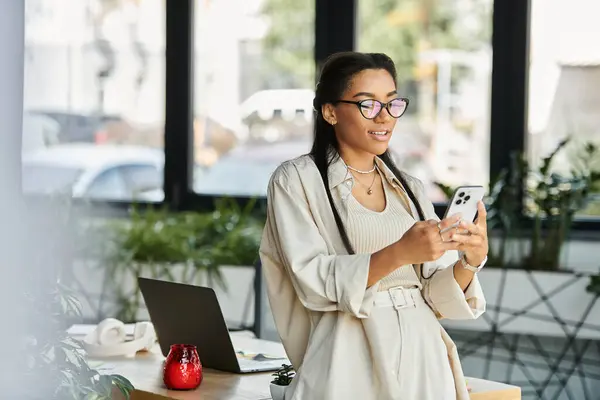 A beautiful young woman stands in an office, happily using her smartphone while working. — Stock Photo
