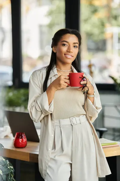 Eine selbstbewusste junge Frau steht in ihrem stilvollen Büro und hält lächelnd einen knallroten Becher in der Hand.. — Stockfoto