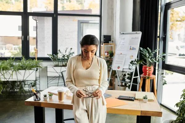 A focused young woman stands at her desk, contemplating her work while surrounded by greenery. — Stock Photo