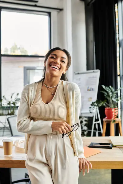 Uma jovem mulher fica confiante em um escritório brilhante, sorrindo e segurando óculos, irradiando alegria. — Fotografia de Stock