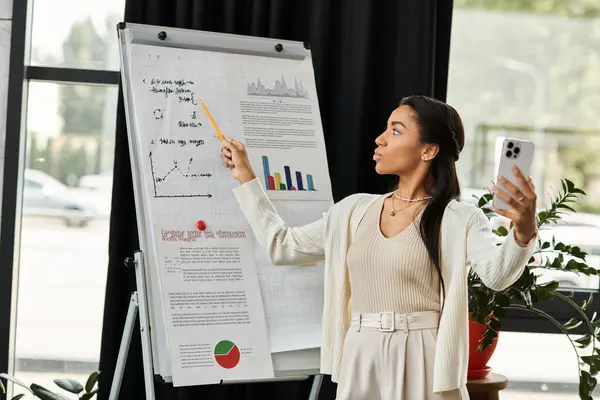 Una giovane donna spiega energicamente le tendenze dei dati mentre tiene lo smartphone in un ufficio. — Foto stock