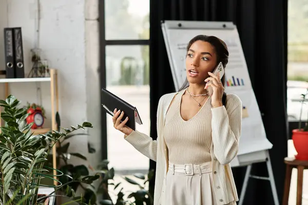 A young woman multitasks in a bright office space, talking on her phone and holding a tablet. — Stock Photo