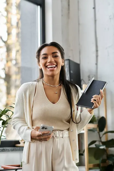 Une jeune femme en tenue stylée s'engage joyeusement tout en tenant un carnet et un téléphone dans son bureau. — Photo de stock