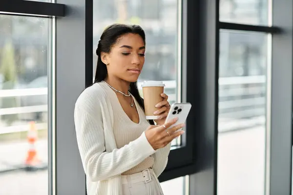 In a modern office, a young woman sips coffee while focused on her smartphone. — Stock Photo
