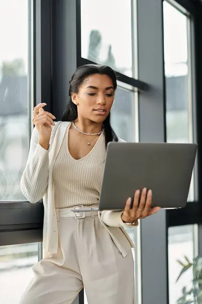 A young, beautiful woman attentively reviews her laptop while leaning against a window. — Stock Photo