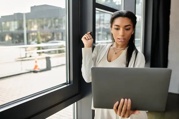 A young woman concentrates on her laptop while standing by a window, engaged in work. — Stock Photo