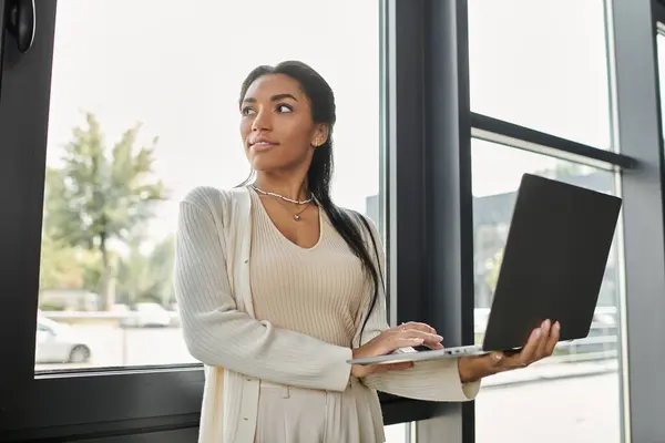 Una joven confiada se para junto a una ventana, interactuando con su portátil en una oficina moderna. - foto de stock