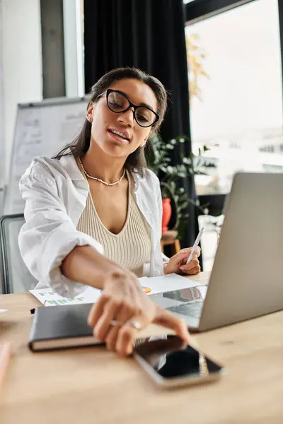 Une jeune femme concentrée analyse des données sur son ordinateur portable tout en cherchant son téléphone. — Stock Photo