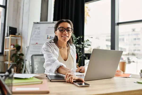 A young woman in a stylish office smiles while working on her laptop and organizing documents. — Stock Photo