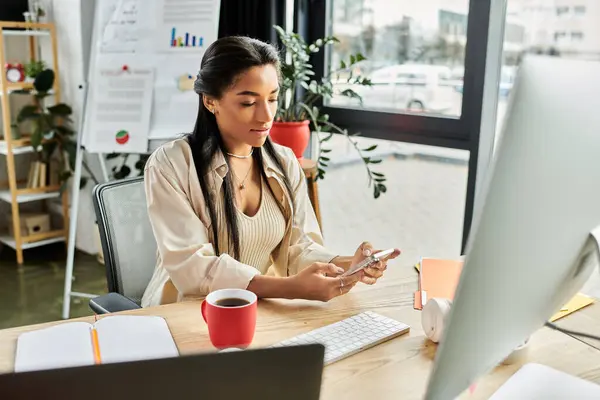 Eine junge Frau in einem schicken Büro checkt ihr Smartphone und genießt eine warme Tasse Kaffee. — Stockfoto