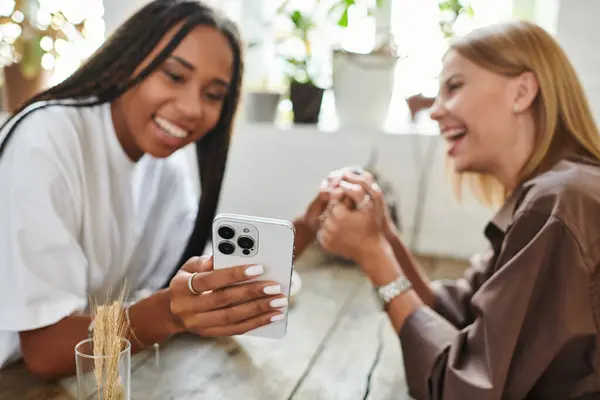 Dos mujeres se relajan en un café, compartiendo un momento alegre mientras miran un teléfono juntas, sonriendo brillantemente. - foto de stock