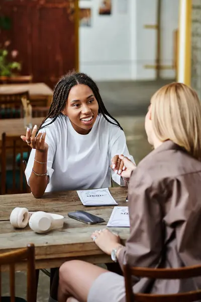A cheerful African American woman with braided hair smiles warmly at her girlfriend during a cafe date. — Stock Photo
