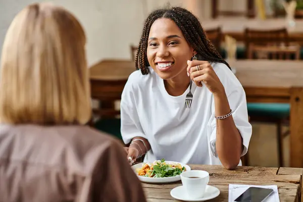 A joyful brunch unfolds as a smiling woman with braided hair shares moments with her girlfriend at a cafe. — Stock Photo