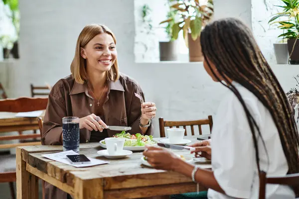 In einem warmen Café teilt eine lächelnde Frau eine Mahlzeit mit ihrer Freundin, umgeben von viel Grün und Liebe. — Stockfoto