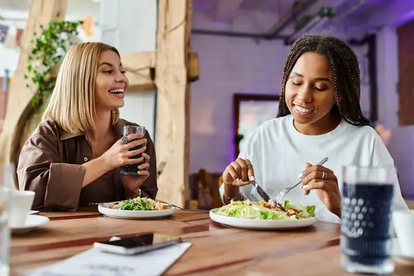 A smiling woman with braided hair enjoys a meal while her girlfriend looks on with delight at a cafe. — Stock Photo