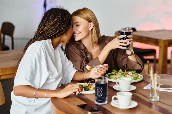A joyful African American woman smiles at her girlfriend in a warm cafe as they savor their food and drinks. — Stock Photo