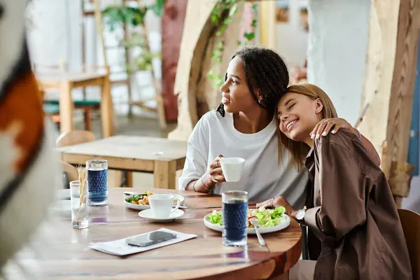 A multicultural lesbian couple smiles together at a cafe, enjoying breakfast and each others company. — Stock Photo