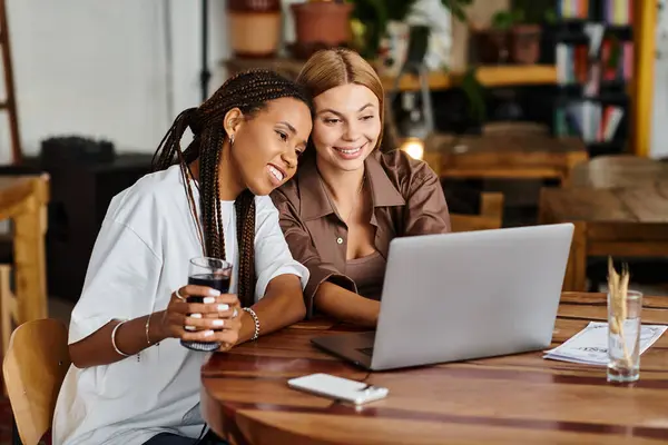 Zwei fröhliche Frauen sitzen zusammen, genießen Gesellschaft und einen Laptop, umgeben von einer lebendigen Café-Atmosphäre. — Stockfoto