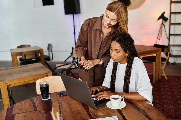 In a welcoming cafe, a smiling woman admires her girlfriend as she works on a laptop, sipping coffee. — Stock Photo