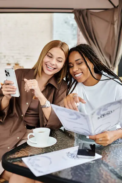 A joyful multiethnic lesbian couple sips coffee and laughs together while browsing a cafe menu. — Stock Photo