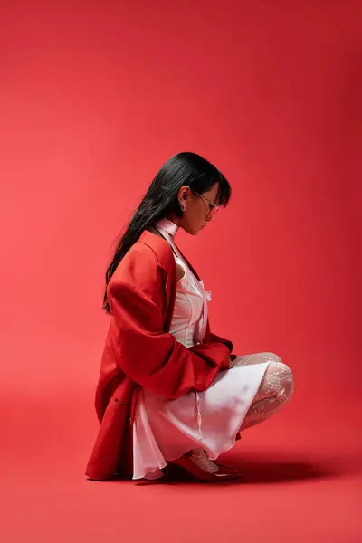A graceful Asian woman kneels, highlighting her vibrant red attire against a bold backdrop. — Stock Photo