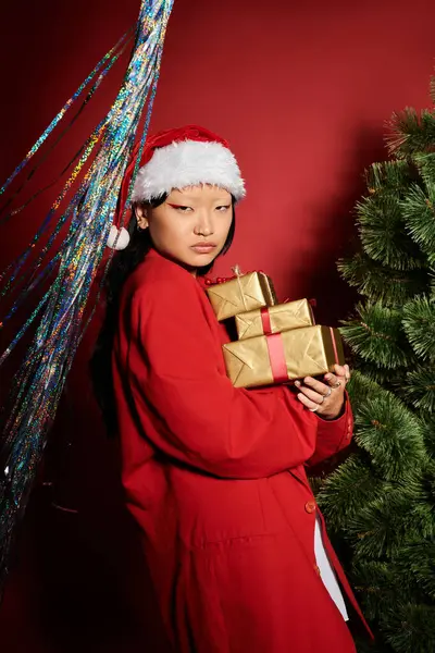 Una donna gioiosa in un cappello di Babbo Natale abbraccia regali colorati vicino a un albero di Natale e decorazioni lucide. — Foto stock