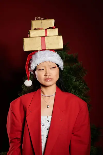 A stylish woman wearing a Christmas hat poses with gift boxes balancing on her head near a tree. — Stock Photo