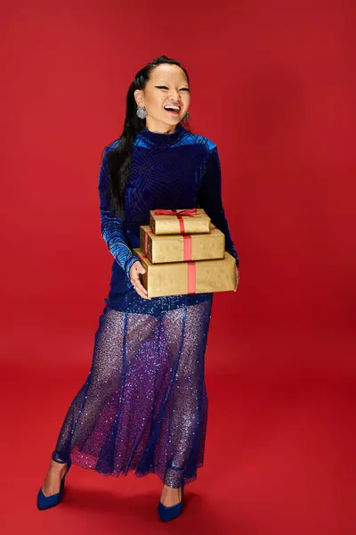 A cheerful Asian woman dressed in a sparkling blue outfit joyfully holds wrapped gifts. — Stock Photo