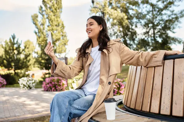 A cheerful young woman dressed warmly sits on a bench, capturing the beauty of fall with her phone. — Stock Photo