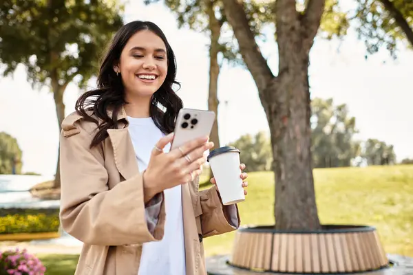 A joyful young woman dressed in cozy autumn attire checks her phone with a warm beverage nearby. — Stock Photo