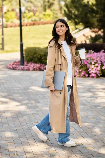 A joyful girl strolls through a vibrant park in autumn, sipping coffee and carrying her laptop. — Stock Photo