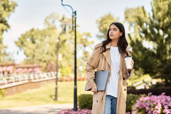 A beautiful young woman in autumn attire walks through a lush park, holding coffee and laptop. — Stock Photo