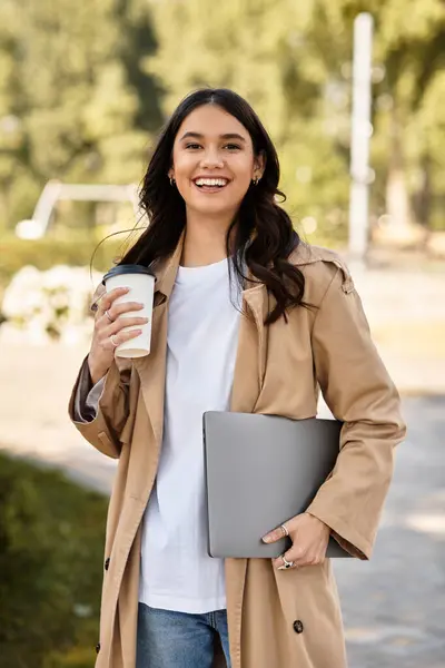 A young woman smiles, strolling through a vibrant park with warm coffee, dressed for autumn. — Stock Photo