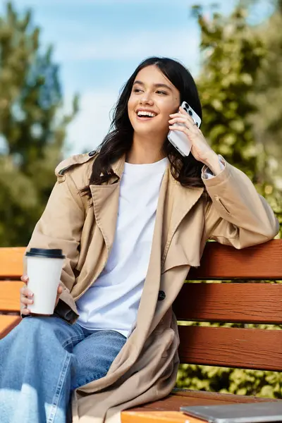 A young woman in warm attire smiles while chatting on the phone, sipping coffee in the park. — Stock Photo