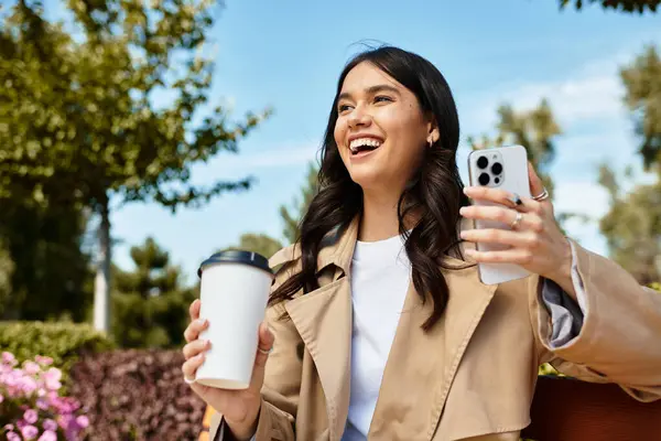 Uma jovem mulher em trajes de outono sorri com café e seu telefone ao ar livre. — Fotografia de Stock